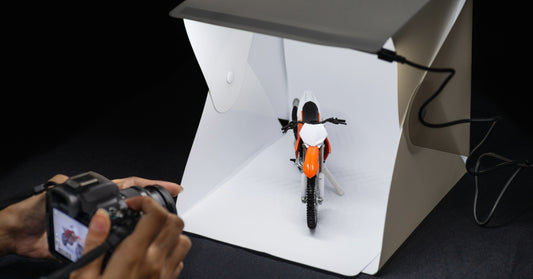 A picture of a mini motorcycle in a lightbox with someone's hands holding a camera taking a picture of it. Or maybe it's a regular sized motorcycle in a super big lightbox and some giant's hands. Who knows.